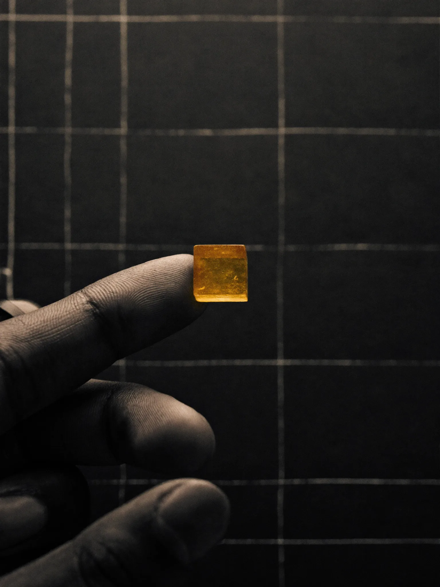 Editorial scene: an artisan's fingertip holding a single glowing golden grain over a dark drafting grid